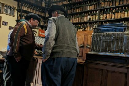 Two men in traditional dress standing at a bar with bottles in the background - San Antonio de Areco