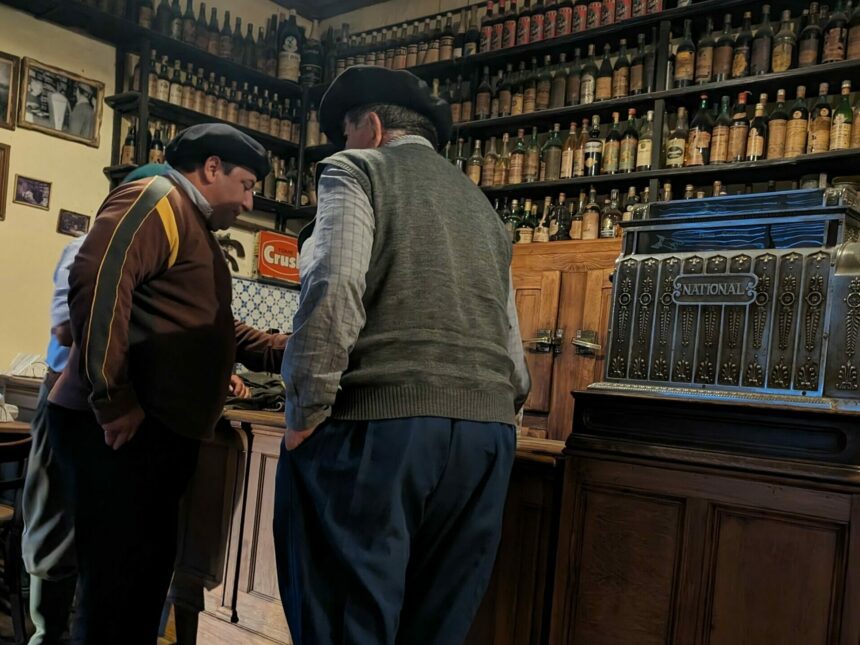 Two men in traditional dress standing at a bar with bottles in the background - San Antonio de Areco