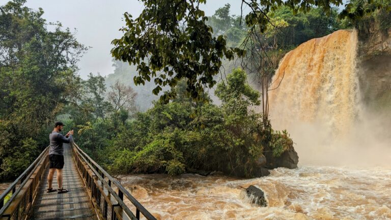 A man standing on a bridge taking a photo of a large waterfall - Iguazu Falls from the Argentina side