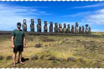 A man standing in front of a line of Easter Island heads behind a grassy field