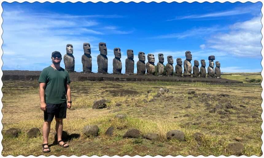 A man standing in front of a line of Easter Island heads behind a grassy field