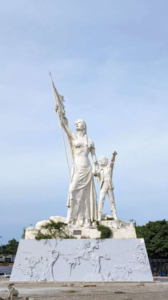 A white statue of a woman holding a torn flag and a child pointing to the sky that is visited during the Circuito de Oro