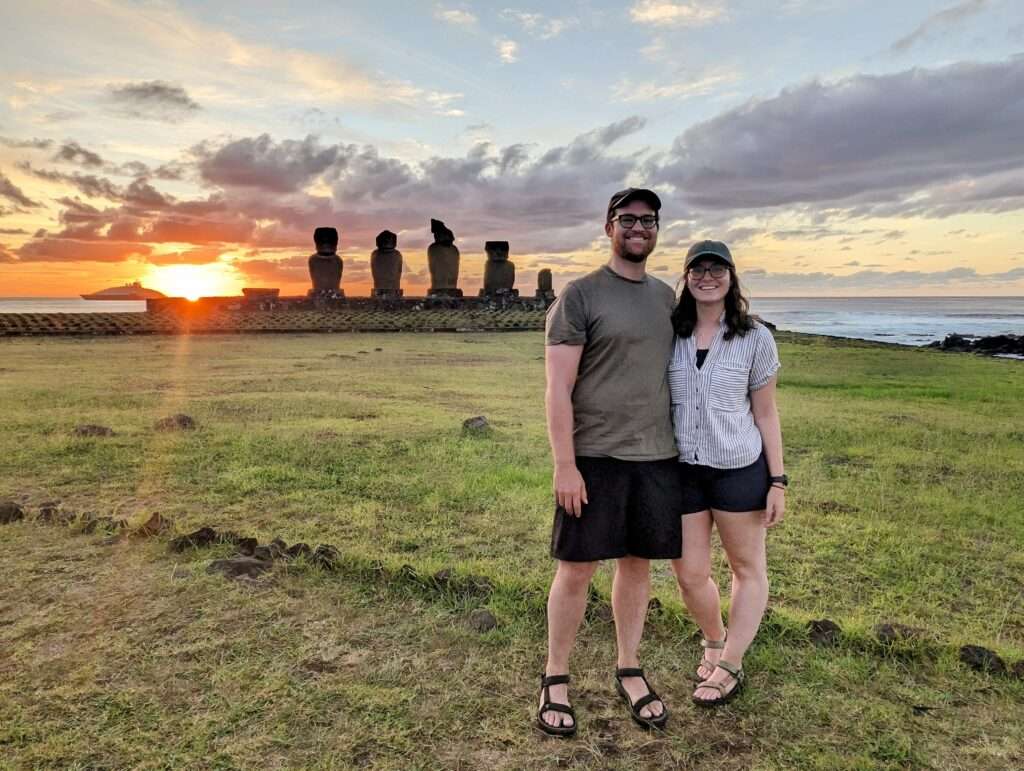 Two people standing in front of a set of statues during a sunset - How to Visit Easter Island