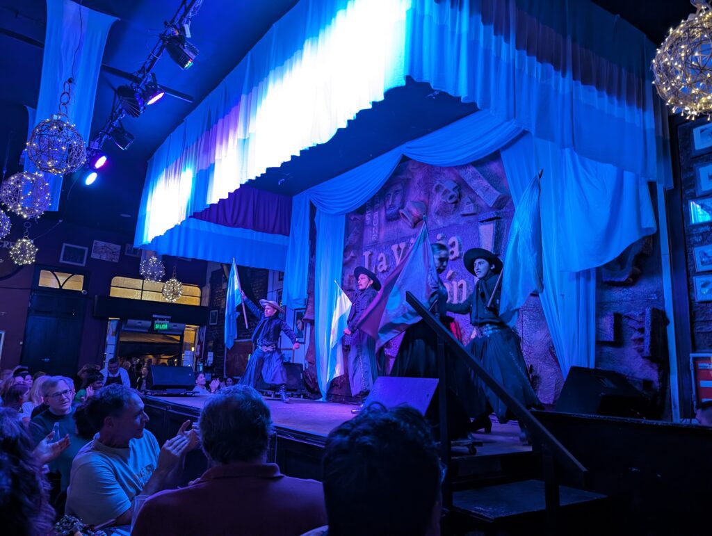 A group of dancers on a stage at a restaurant flooded with blue lights showing traditional dancing when visiting Salta