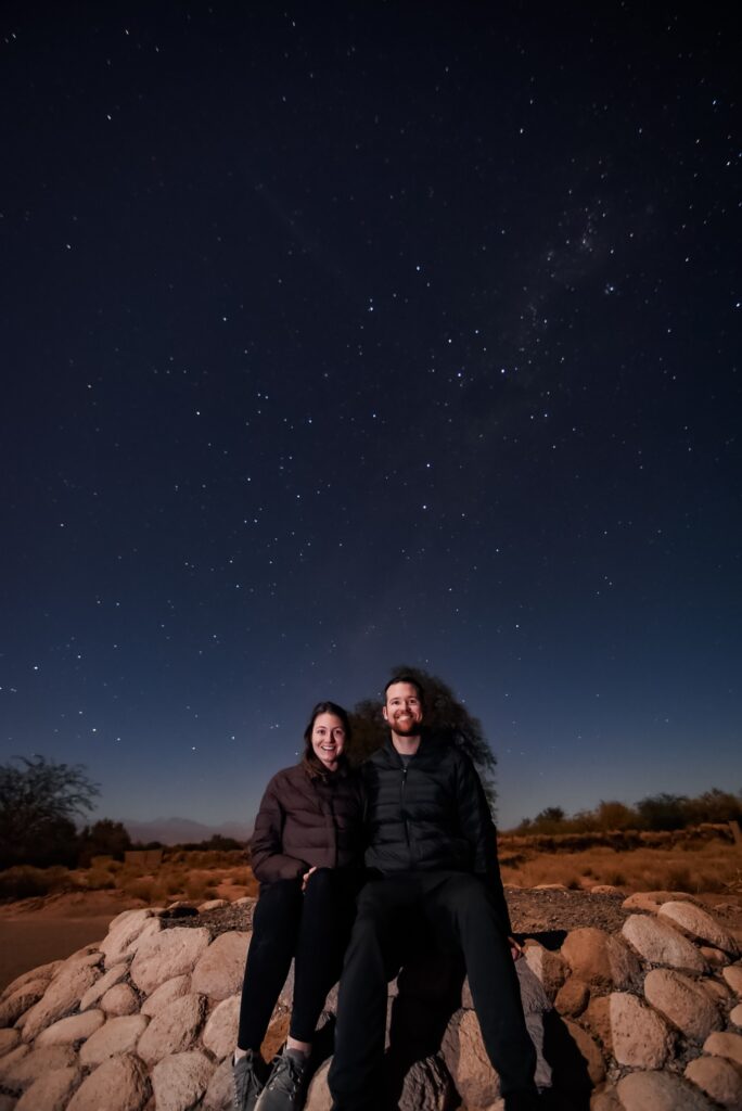 Two people sitting on a rock in front of a starry sky in the Atacama desert
