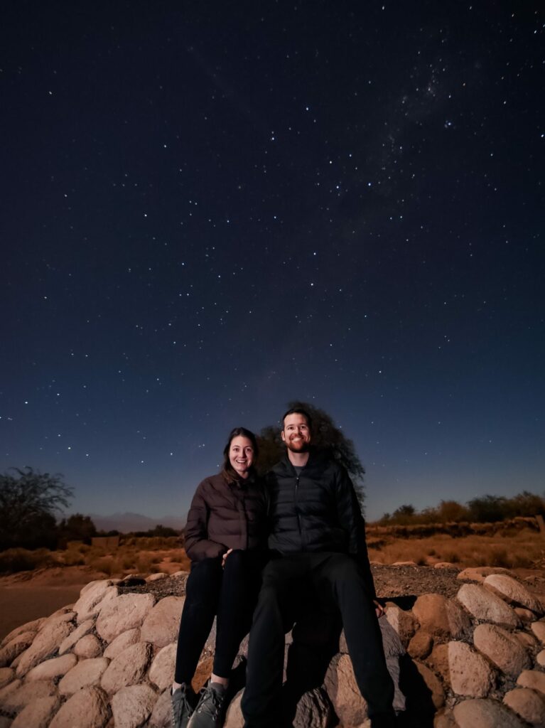 Two people sitting on a rock in front of a starry sky in the Atacama desert