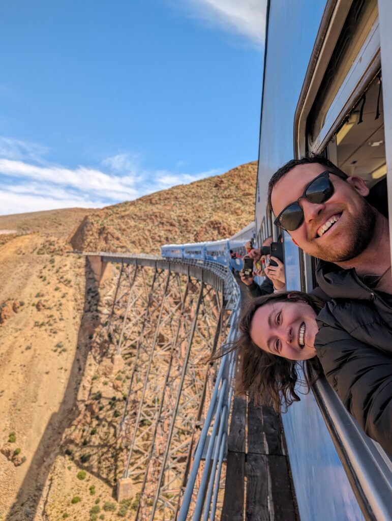 two people leaning out of a blue train on a bridge over a canyon