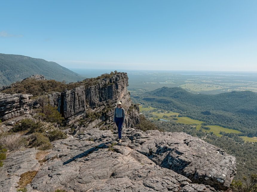 A woman standing in front of a rock at the peak of a small mountain overlooking a green valley - Attractions in Grampians