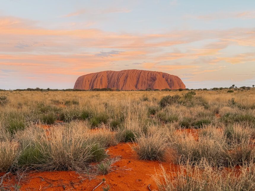 The large rock of Uluru surrounded by brush and red dirt at sunset