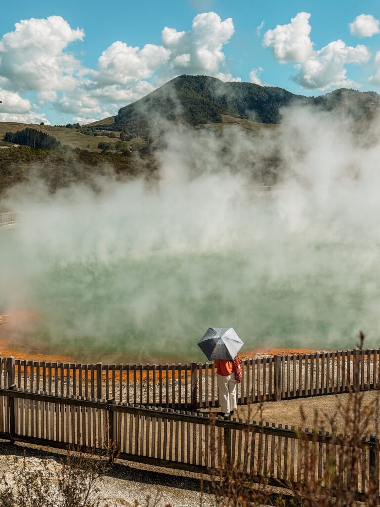 A person with an umbrella standing in front of a turquoise and orange pond with steam rising from it in Rotorua