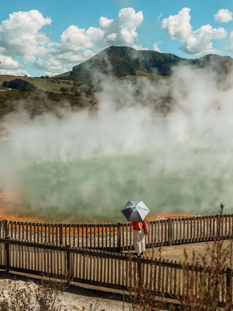 A person with an umbrella standing in front of a turquoise and orange pond with steam rising from it in Rotorua