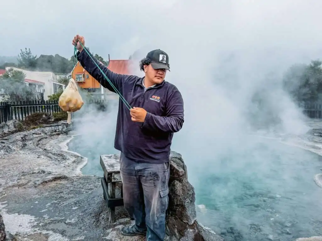 A man holding a bag of corn in front of a steaming pool as part of a Rotorua itinerary