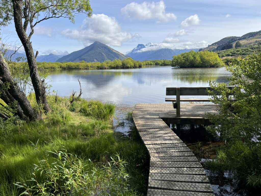 A bench at the edge of a lake on a wooden path with mountains behind when visiting Queenstown