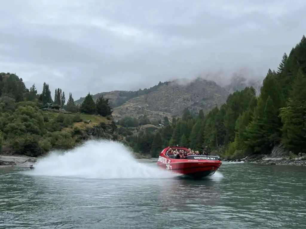 A boat splashing through a river with mountains behind for an activity that can be done when visiting Queenstown