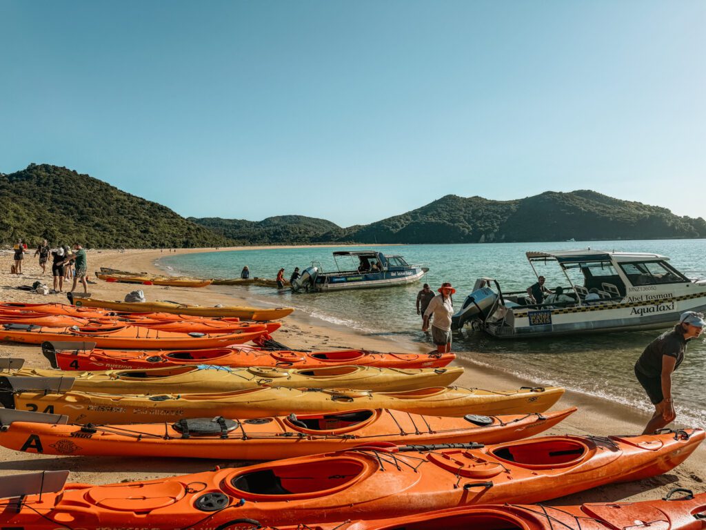A row of kayaks waiting on a beach with people getting out of speed boats docked along the beach in Abel Tasman National Park