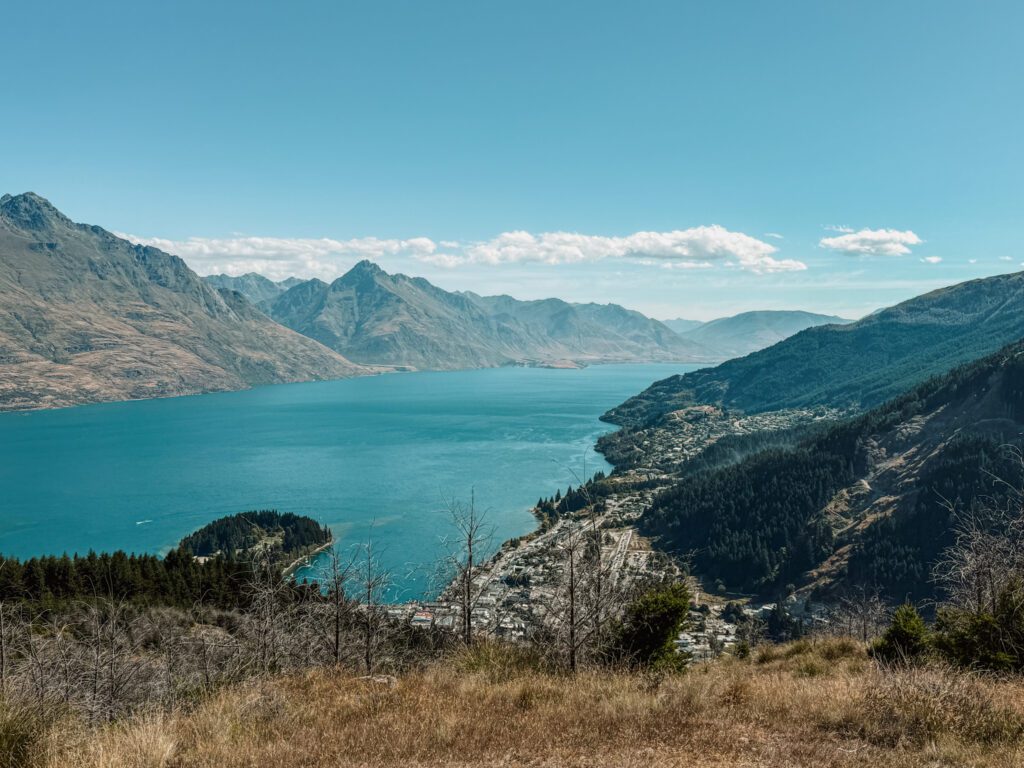 The view of a town at the base of a lake from the top of a hill on an itinerary between Queenstown and Christchurch