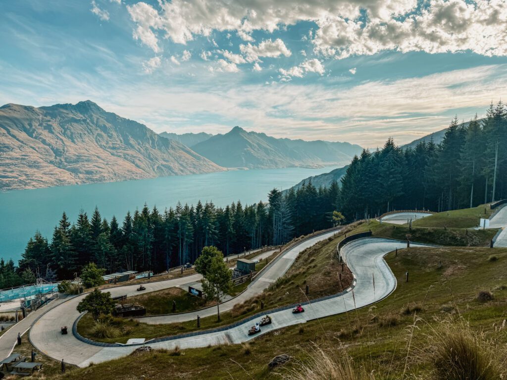 A winding road with small cars on it on a mountain over a lake, something that can be done on the itinerary between Queenstown and Christchurch