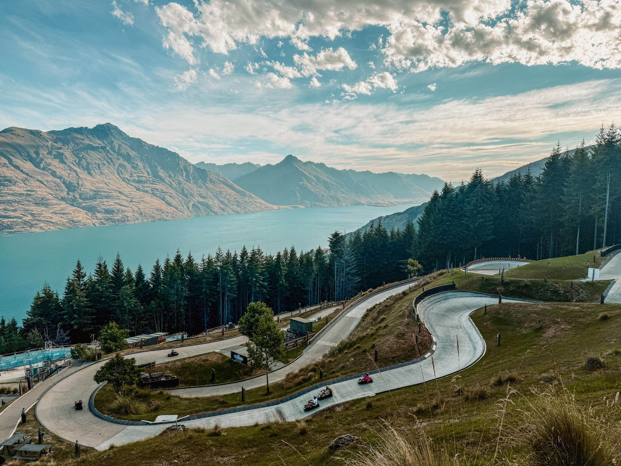 A winding road with small cars on it on a mountain over a lake, something that can be done on the Queenstown itinerary