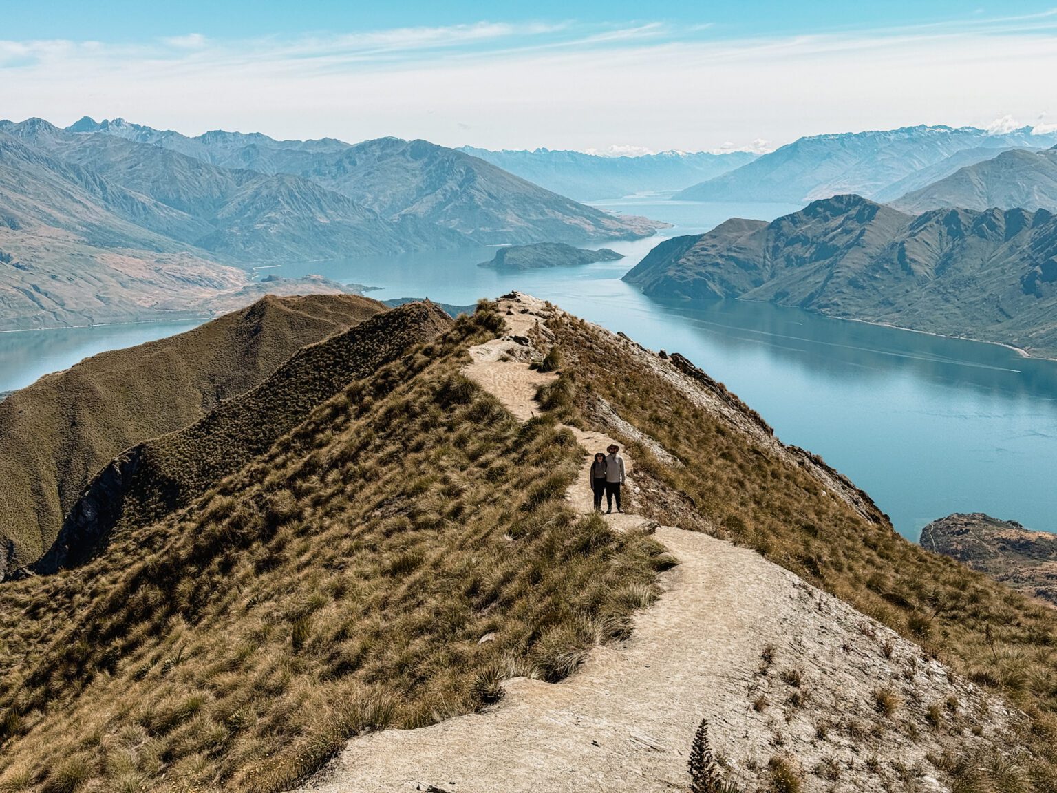 Two people standing on a path on the peak of a mountain overlooking a lake on the Queenstown to Christchurch road trip