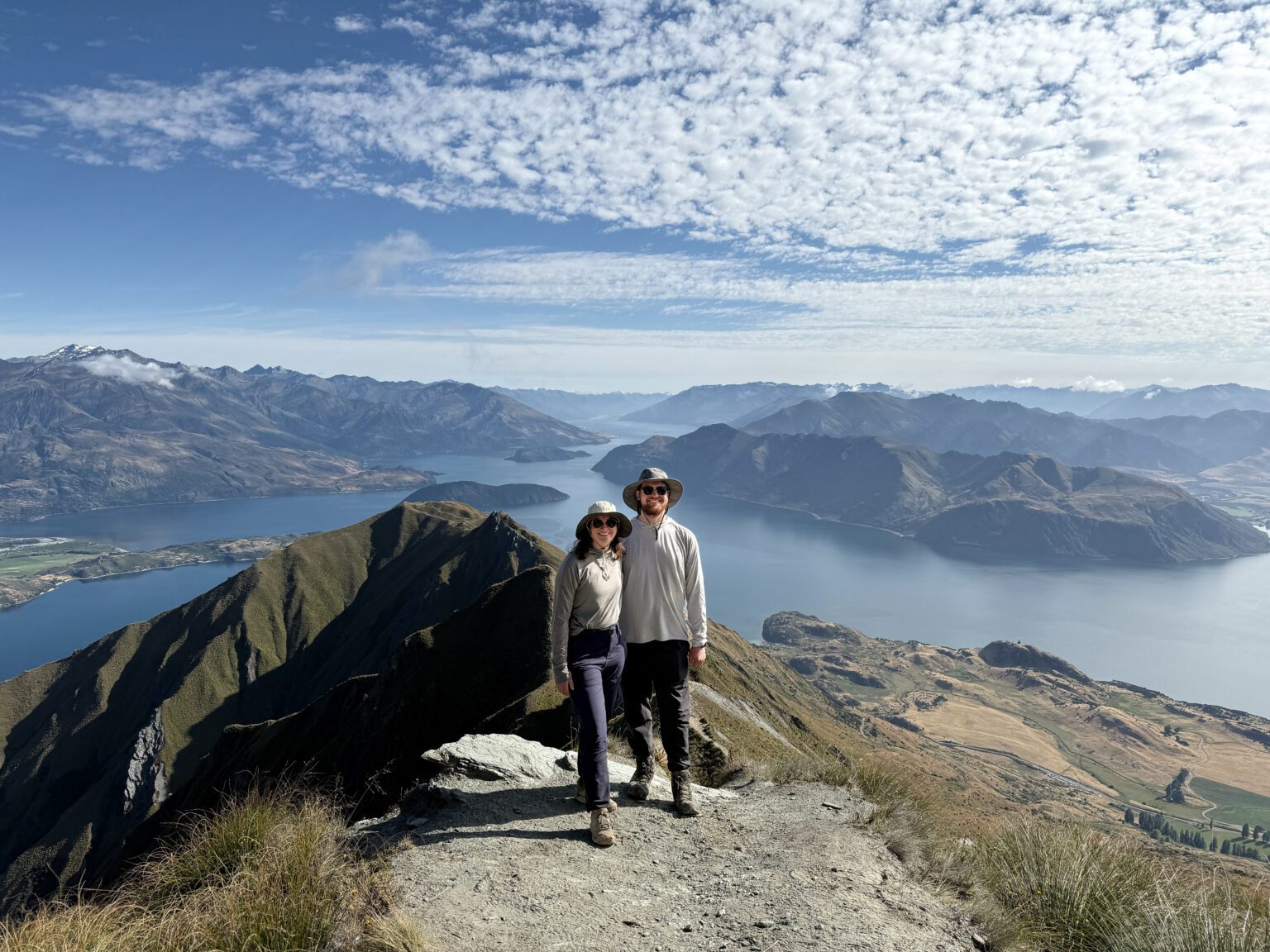 Two people posing on the edge of a mountain overlooking a lake within mountains near the Southern Alps
