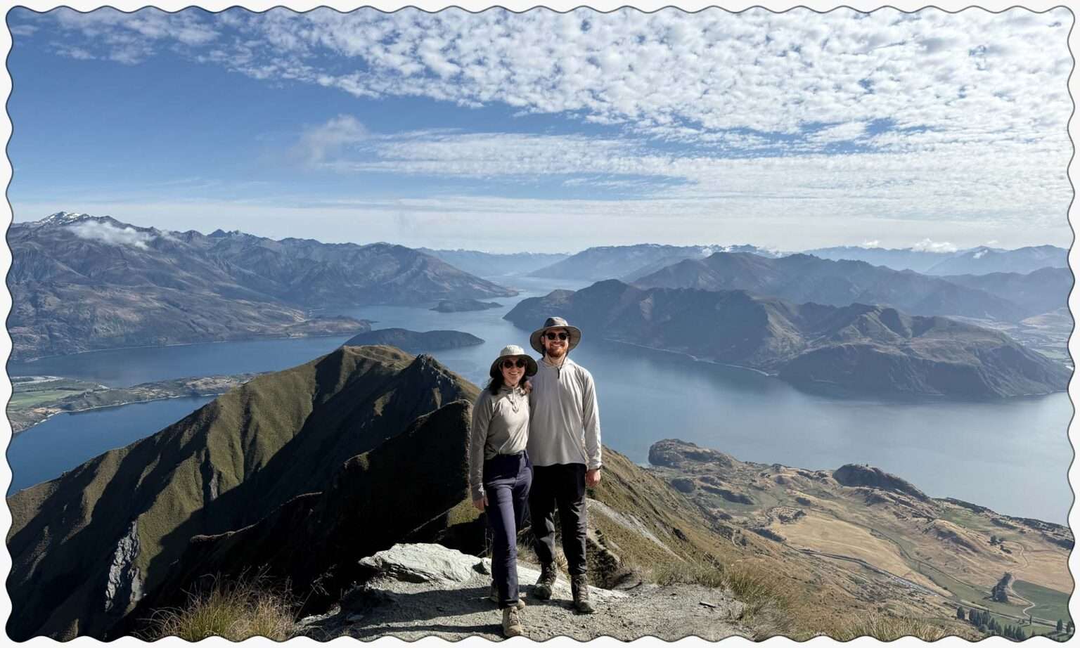 Two people posing on the edge of a mountain overlooking a lake within mountains near the Southern Alps