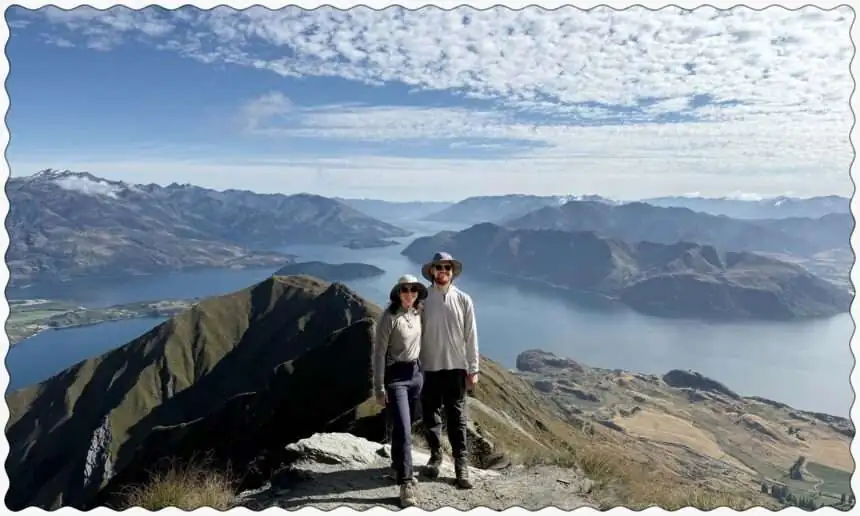 Two people posing on the edge of a mountain overlooking a lake within mountains near the Southern Alps