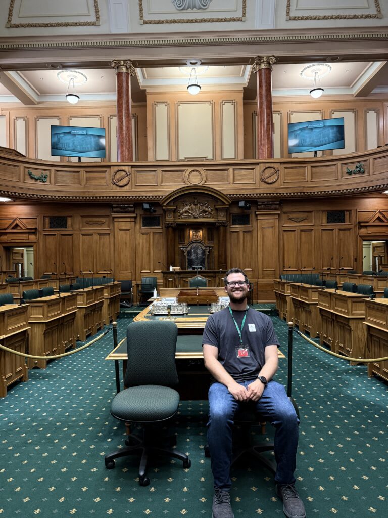 A man sitting on a chair on the floor of a parliament room with green carpet in New Zealand's Capital city