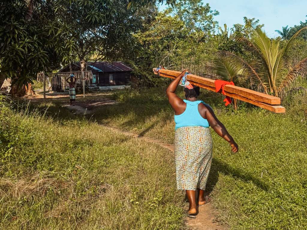 A woman walking on a small path through a village carrying large 2x4 pieces of wood on her head