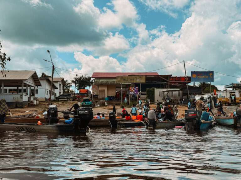 A dock of pirogues on the side of a river with buildings in the background - Guianas Itinerary