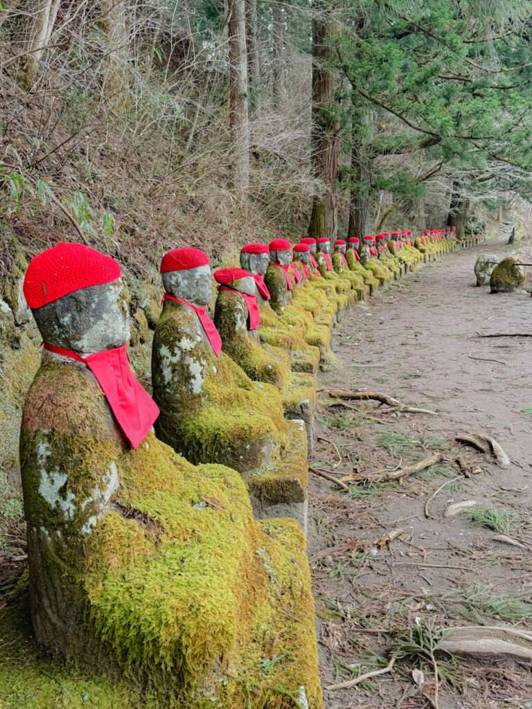 A row a stone statues with moss growing on them with red hats and bibs along a forest pathway in Nikko