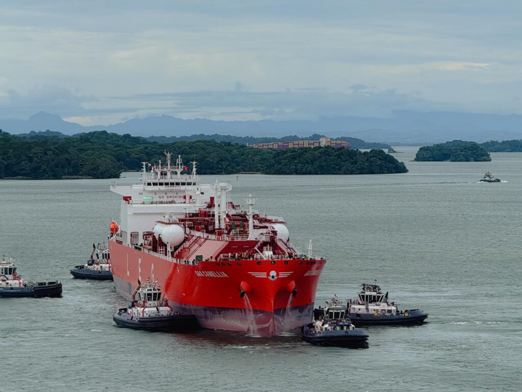 A red LNG tanker surrounded by tugboats approaching the new Agua Clara locks viewed from the elevated visitor center viewpoints, with Gatun Lake, jungle islands, and mountains in the background, Panama Canal