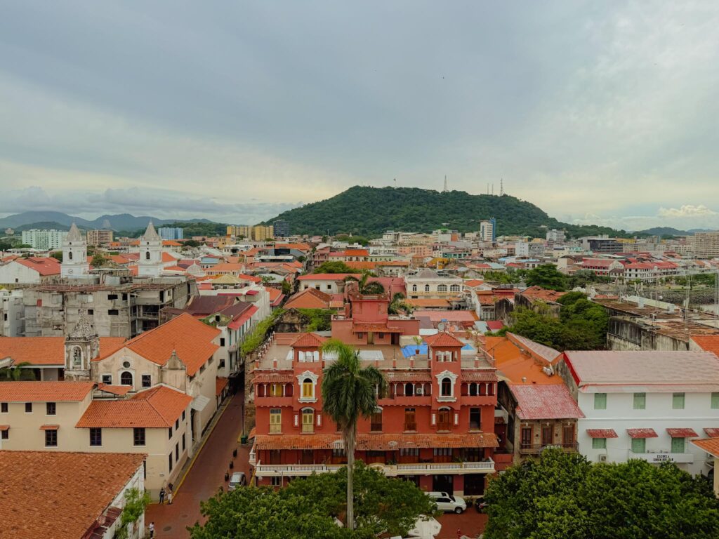 Aerial view over the red-tiled rooftops of Casco Viejo in Panama City, with a green hill and the surrounding city visible in the background