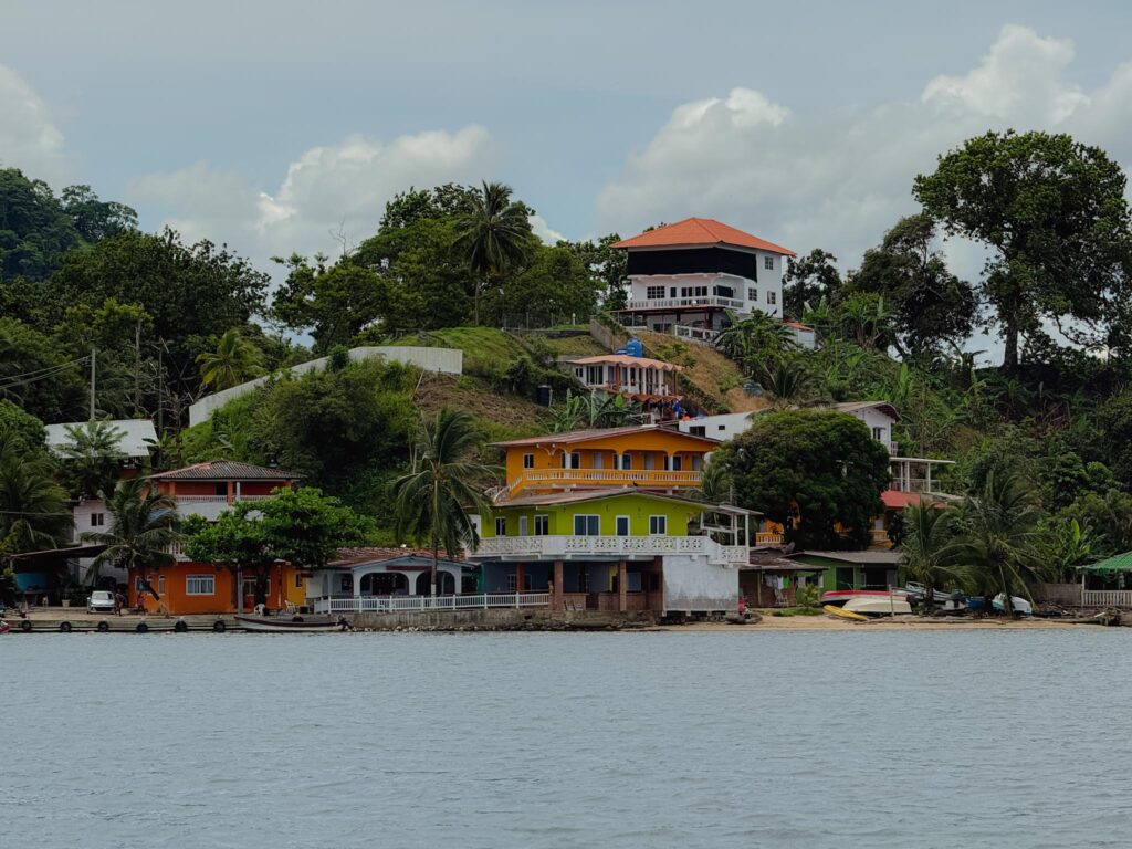 Colourful buildings stacked on a hillside in Portobelo, Panama, viewed from the water, with palm trees and small boats along the Caribbean coastline