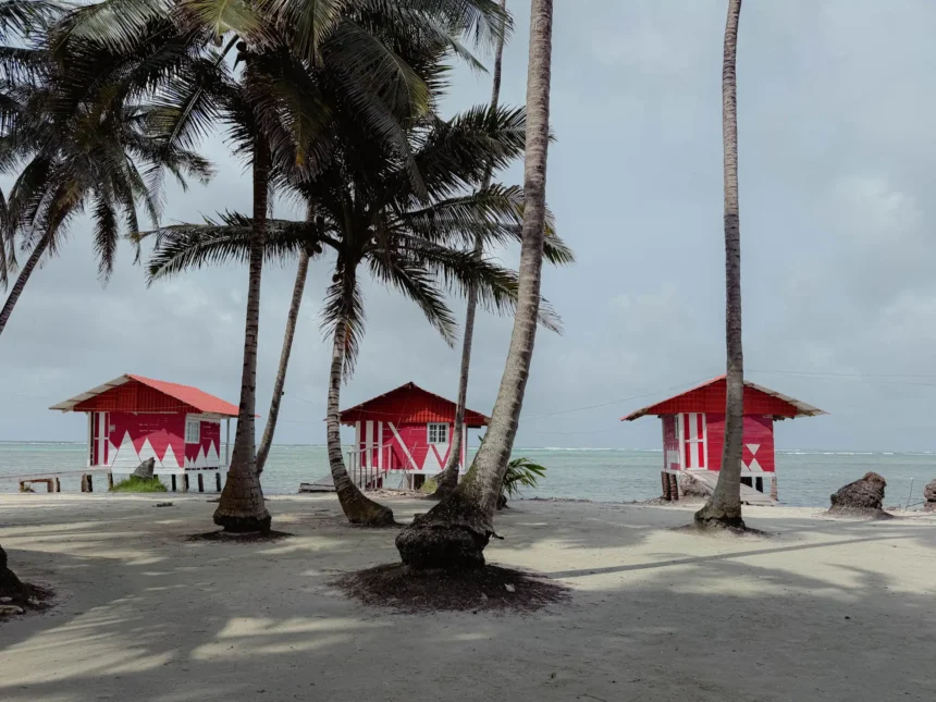 Three red painted wooden Guna-run island cabins between palm trees with the Caribbean Sea behind - where to stay San Blas Islands