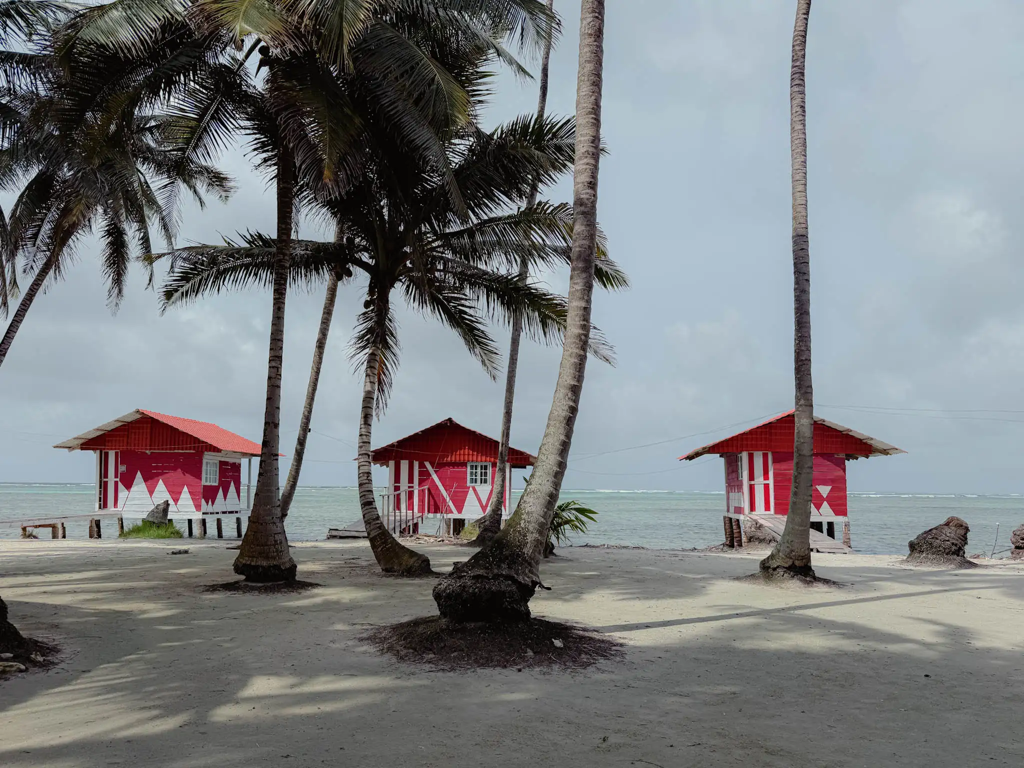 Three red painted wooden Guna-run island cabins between palm trees with the Caribbean Sea behind - where to stay San Blas Islands