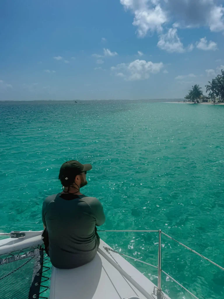 A person sits on the bow of a catamaran looking out at brilliant turquoise water toward a palm-covered San Blas island in the distance -- sailing San Blas Islands Panama