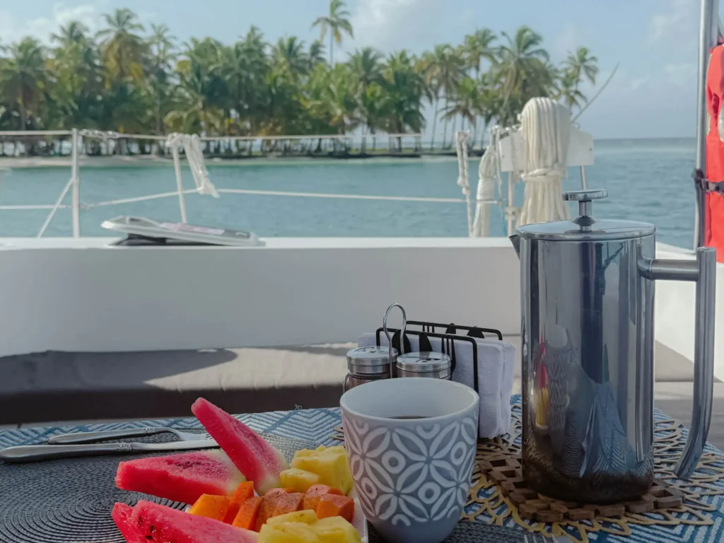 Tropical fruit and a French press on a table on the deck of a catamaran with a palm-covered island visible in the background -- catamaran deck life San Blas Islands