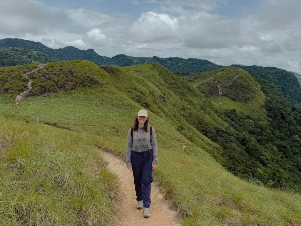 Hiker in sun shirt, hiking pants, cap and backpack walking an exposed ridge trail above Valle de Anton with the crater mountains behind her - Valle de Anton hiking