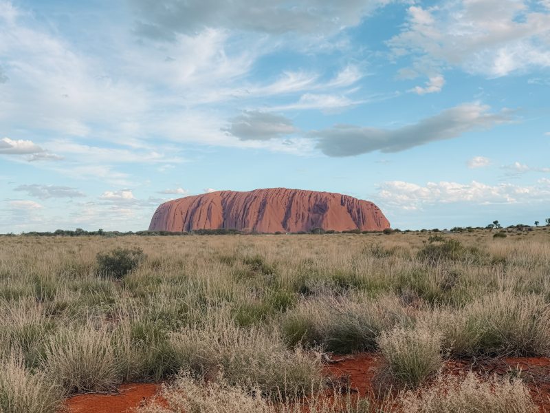 The large rock of Uluru as seen behind low brush and the red dirt landscape