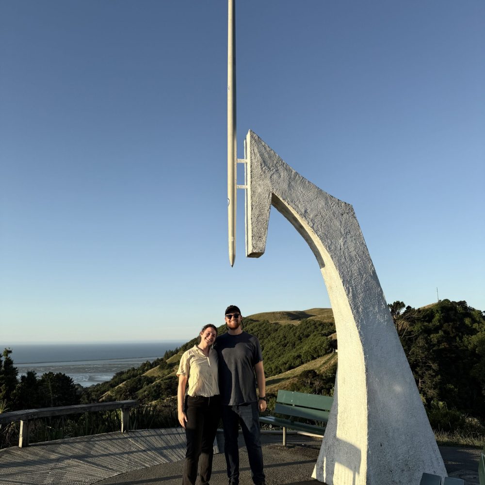 Two people standing under a large monument that looks like the point of a drawing compass on the top of a hill