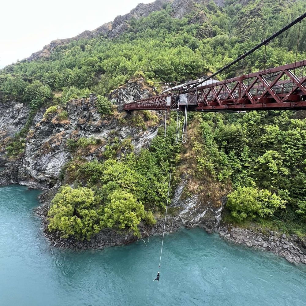 A bridge from a hill covered in plants over a river that can be seen when visiting Queenstown