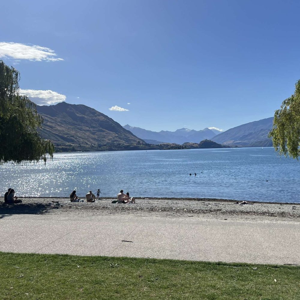 People sitting along a beach on a lake with the Southern Alps mountains in the background