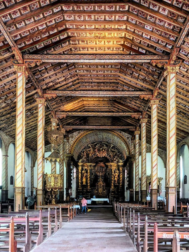 Templo de San Buenaventura – Paraguay – 2 A painted wooden roof and wood pews leading to an ornate altar as part of a church on the Circuito de Oro
