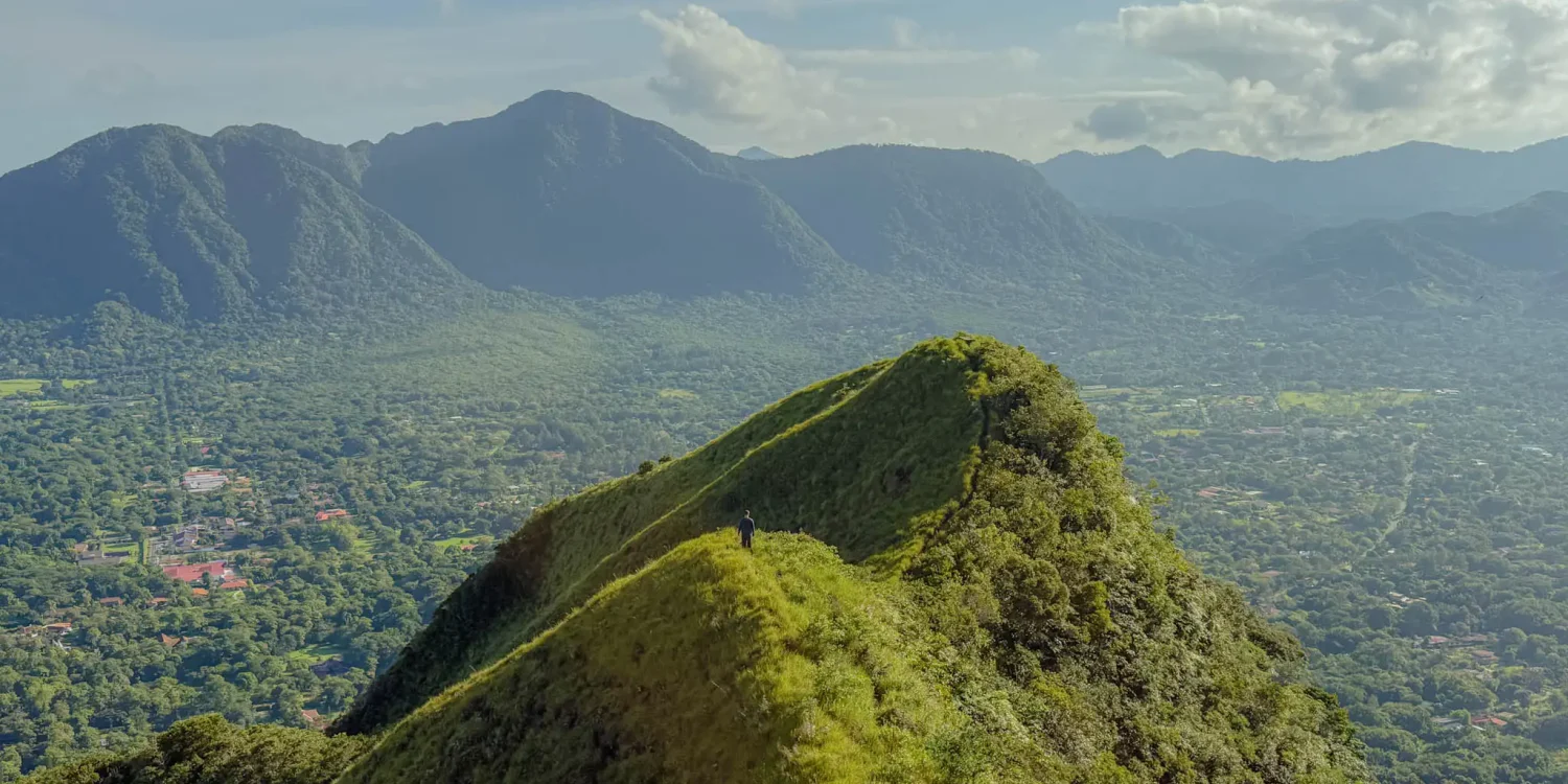 A hiker standing on the narrow Cerro Cara Iguana ridge with the Valle de Anton crater floor and town visible far below -- Valle de Anton Panama